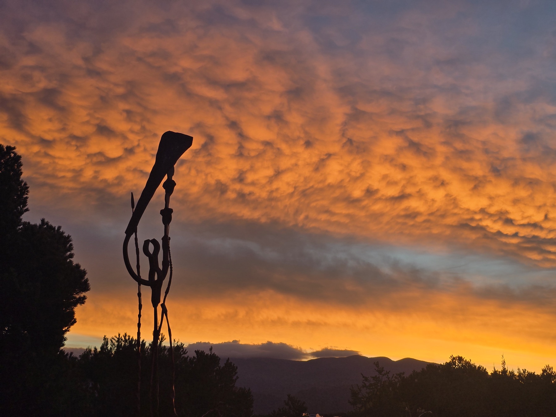 A silhouetted abstract metal sculpture rises against a wavy orange and yellow sunset sky with distant hills below.