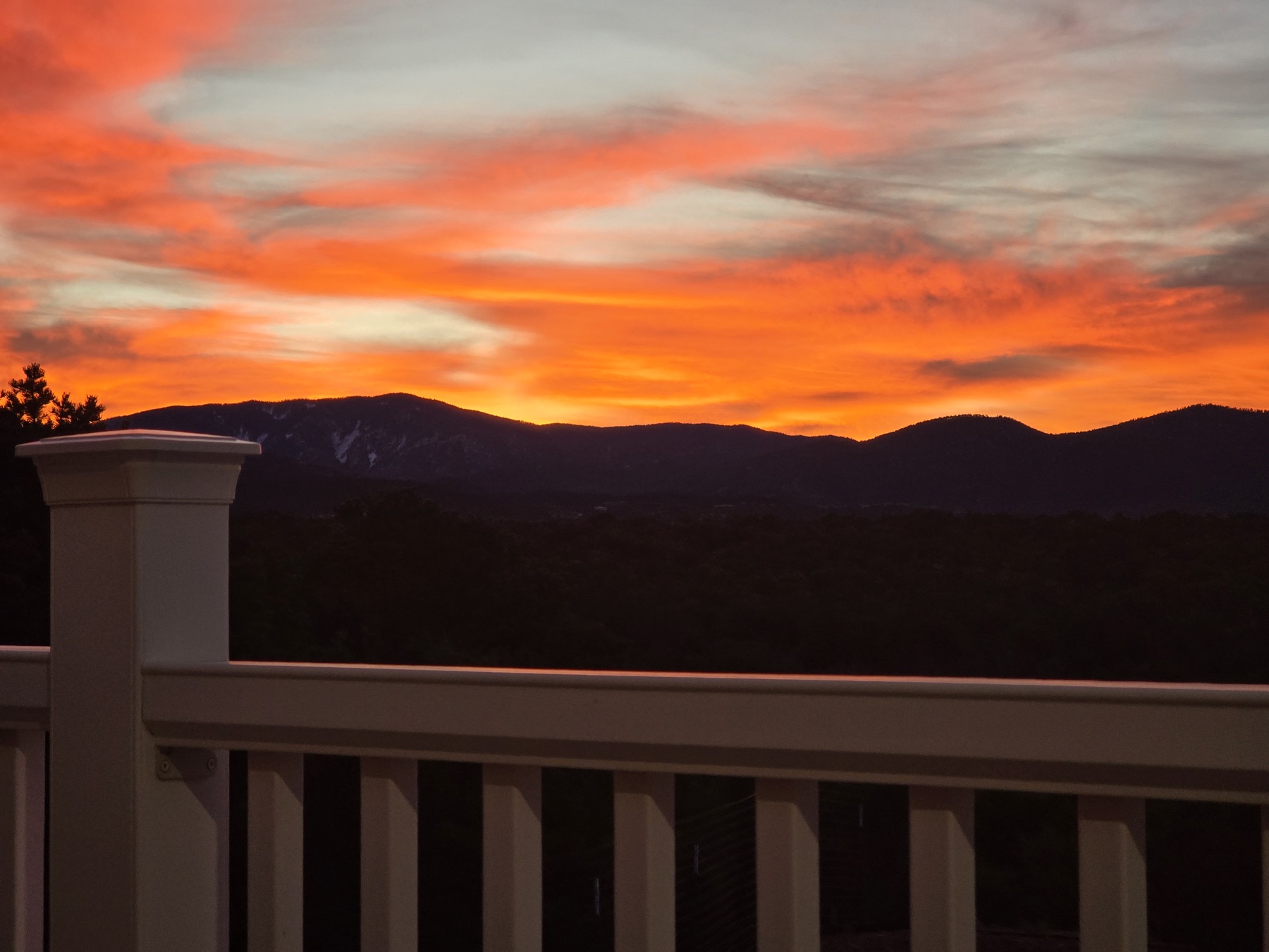 A white wooden balcony railing frames a fiery orange and red sunset sky above dark silhouetted mountain ridges.