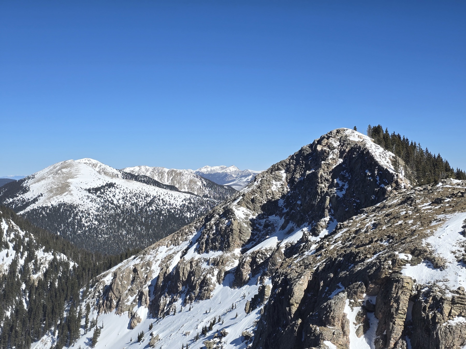 Jagged snow-dusted rocks and evergreens frame a panoramic vista of distant snow-covered mountain ranges under a bright blue sky.
