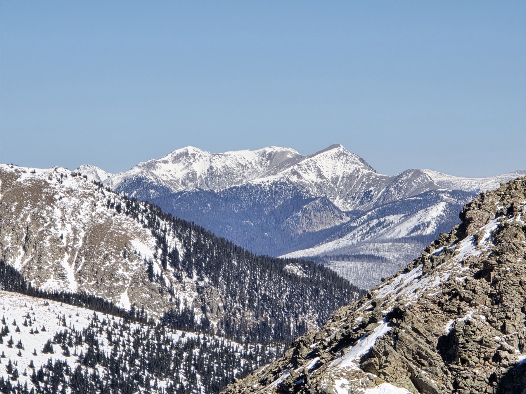 A wide view from a rocky overlook across snow-capped mountain peaks, forested valleys, and a clear blue winter sky.