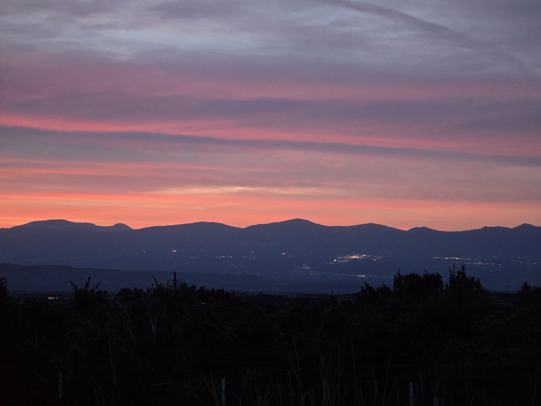 A pink and orange sunset sky hangs over dark mountain silhouettes with clusters of distant valley lights below.