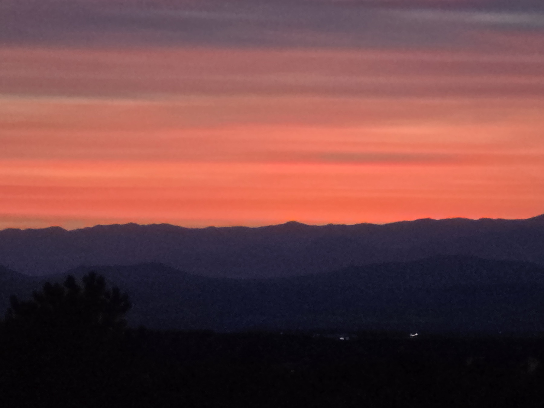 Horizontal stripes of purple, pink, and orange light the sky above dark silhouetted mountain ridges and a foreground tree.