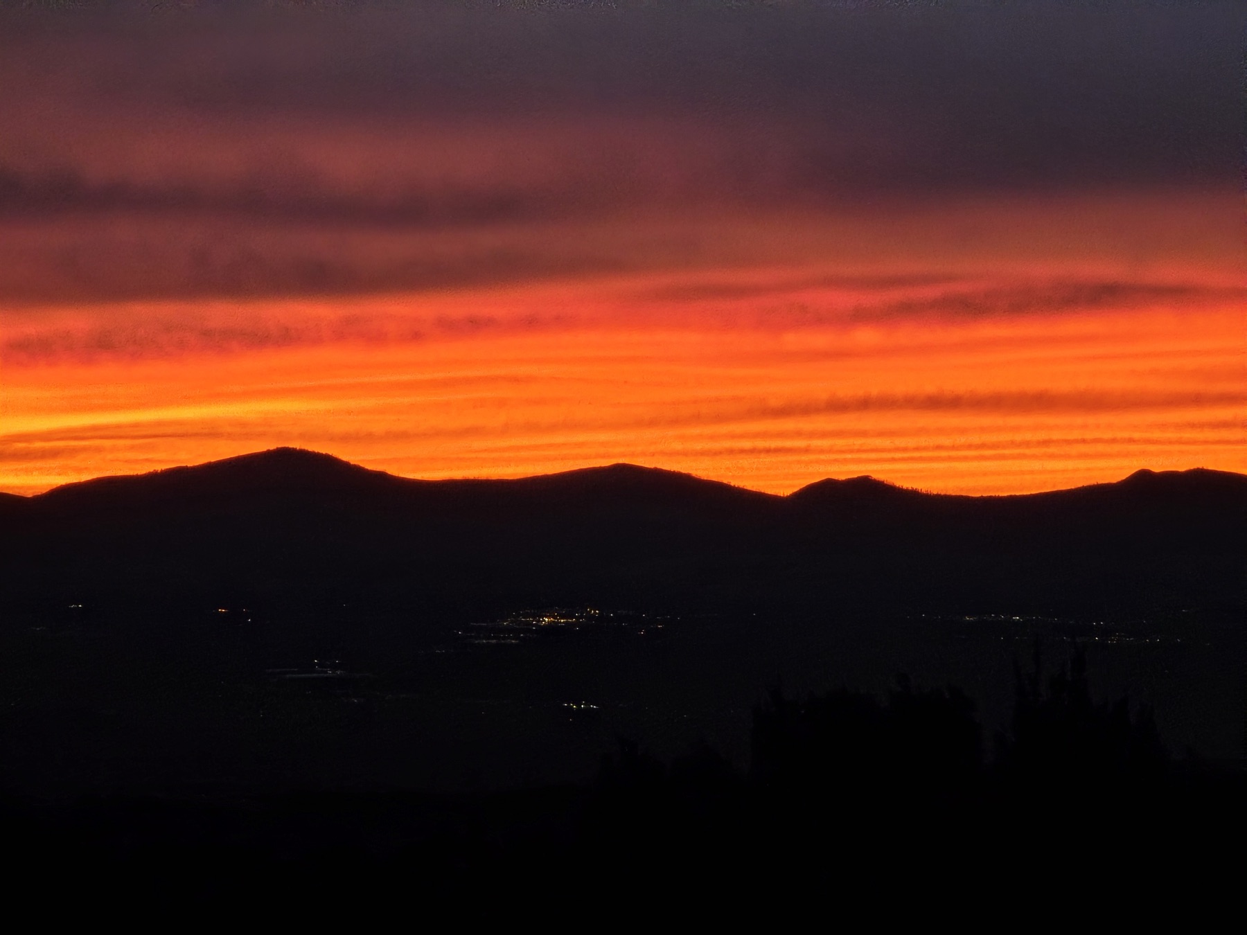 Horizontal bands of fiery orange and red clouds glow above dark mountain silhouettes with scattered lights in the valley below.