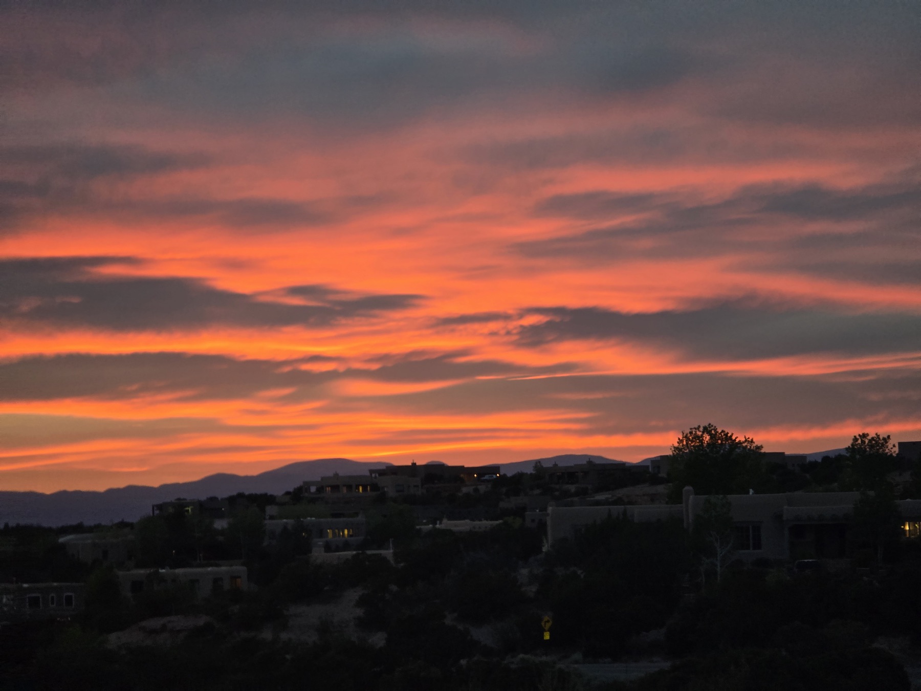 A vivid orange-red sunset with streaky clouds above silhouetted adobe-style homes on a hillside and distant mountains.