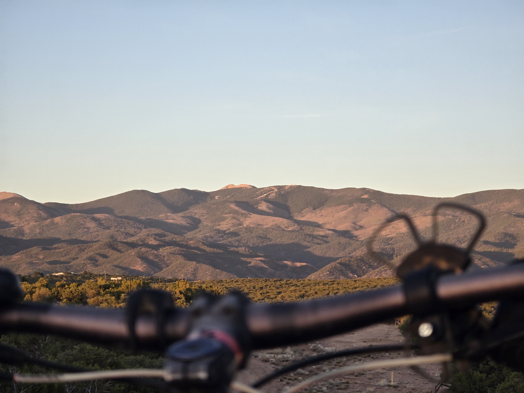 Bicycle handlebars frame a golden-hour view of layered brown mountains and a shrub-covered valley below.
