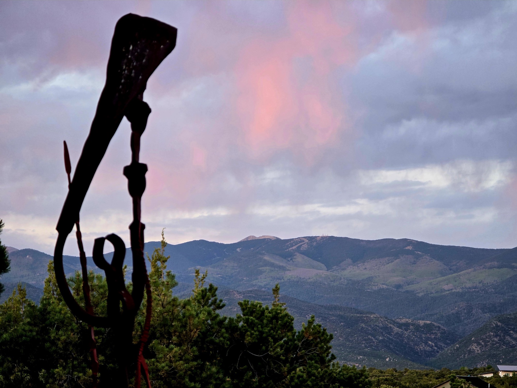 A dark abstract metal sculpture silhouetted against rolling green hills and a pink-purple cloudy sky at sunset.
