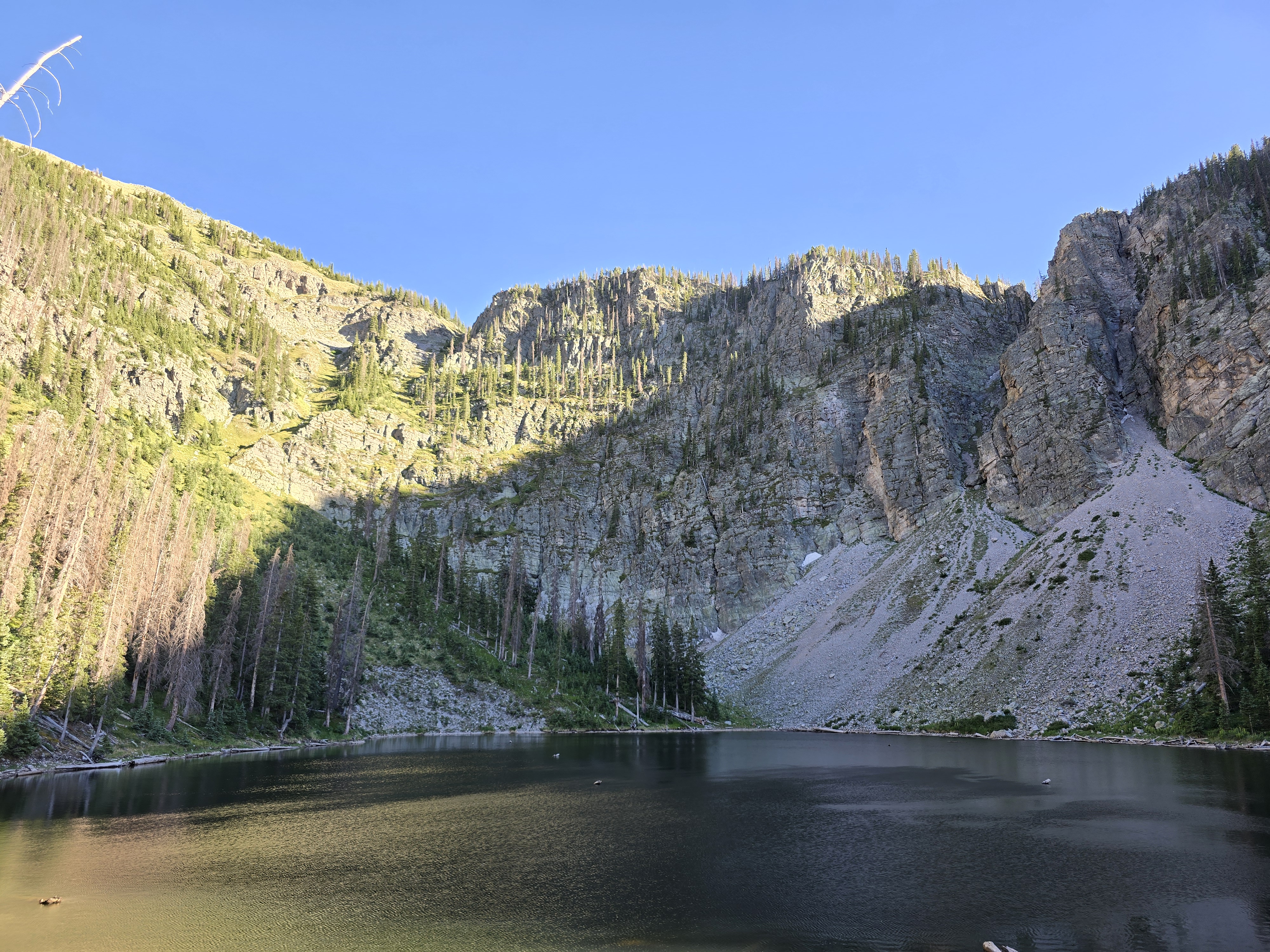 A calm alpine lake reflects steep rocky cliffs and forested slopes beneath a clear blue sky in a high mountain wilderness.