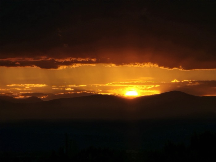 Crepuscular rays stream through dark storm clouds as the sun sets behind silhouetted mountain ridges in golden light.