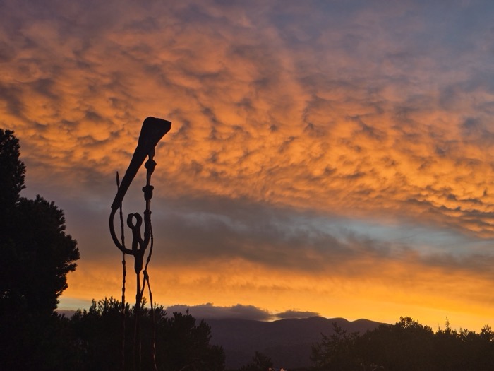 A silhouetted abstract metal sculpture rises against a wavy orange and yellow sunset sky with distant hills below.