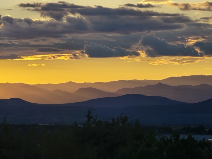 Heavy clouds glow golden yellow above layered purple mountain ridges and dark foreground trees at sunset.