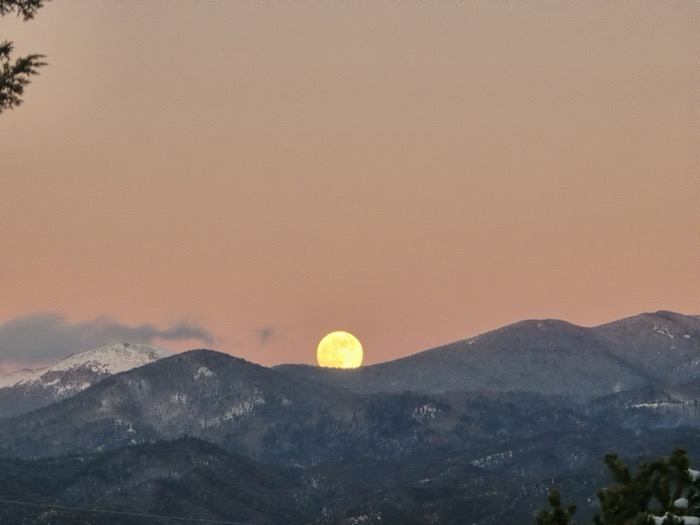 A bright full moon rises above dark snow-patched mountain peaks under a warm peach twilight sky, framed by evergreen branches.