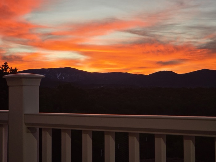 A white wooden balcony railing frames a fiery orange and red sunset sky above dark silhouetted mountain ridges.