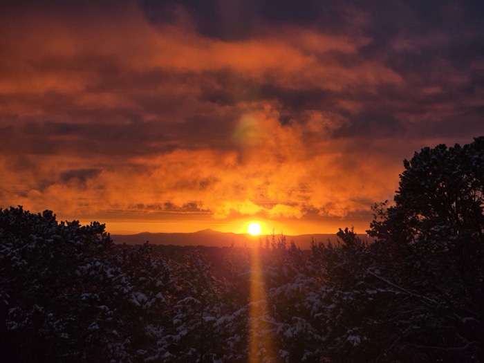 A low winter sun with a golden lens flare shines through billowing orange clouds above snow-covered trees and hills.