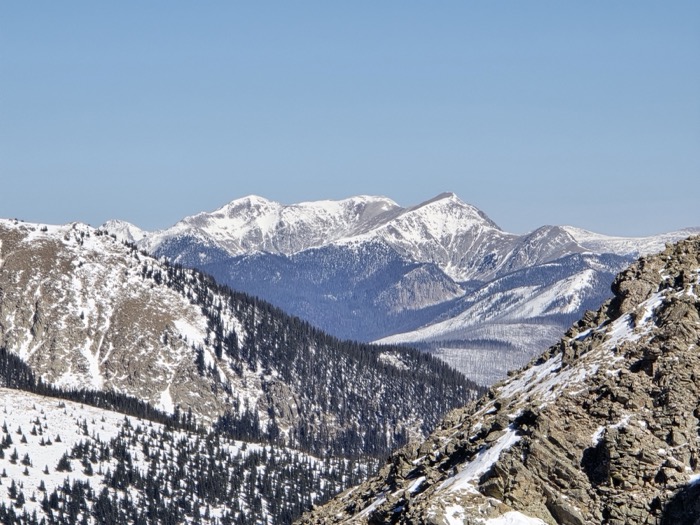 A wide view from a rocky overlook across snow-capped mountain peaks, forested valleys, and a clear blue winter sky.