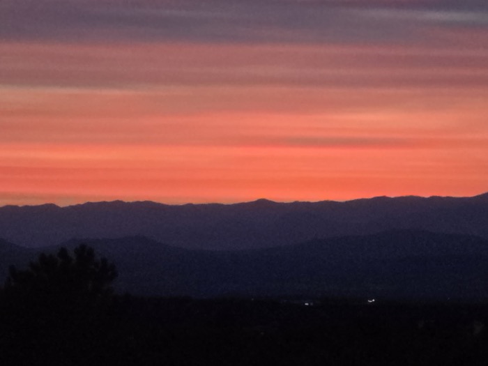 Horizontal stripes of purple, pink, and orange light the sky above dark silhouetted mountain ridges and a foreground tree.