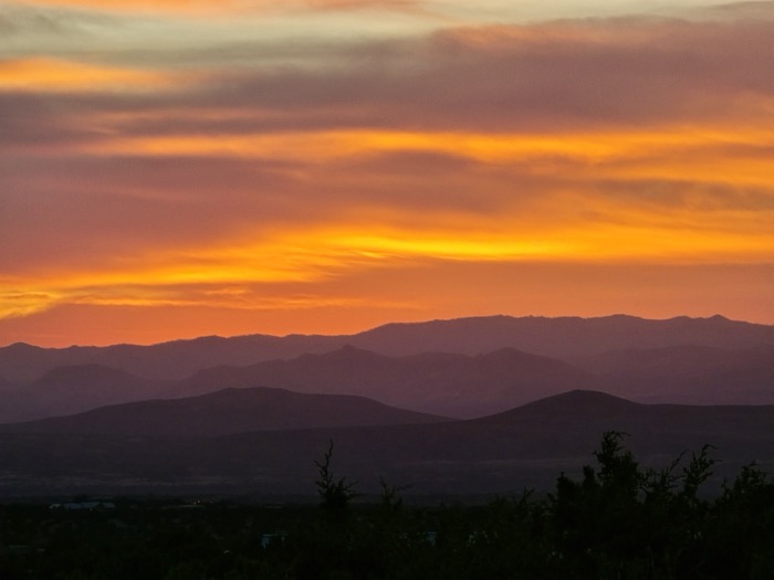 A gradient sky of purple, pink, and orange illuminates layered mountain ridges above dark foreground shrubs.