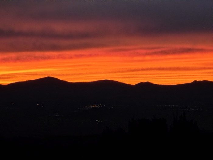 Horizontal bands of fiery orange and red clouds glow above dark mountain silhouettes with scattered lights in the valley below.