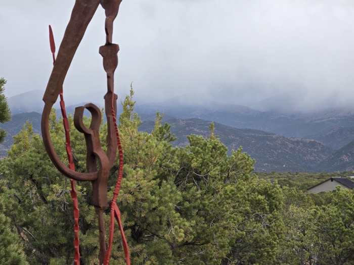 A weathered rusted metal sculpture with red accents stands before fog-shrouded green mountains and pine trees.