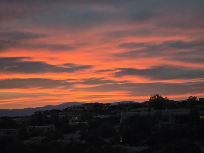 A vivid orange-red sunset with streaky clouds above silhouetted adobe-style homes on a hillside and distant mountains.