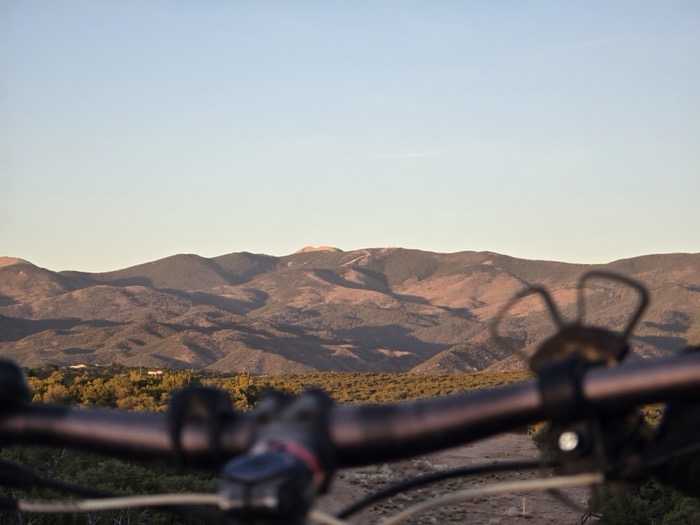 Bicycle handlebars frame a golden-hour view of layered brown mountains and a shrub-covered valley below.