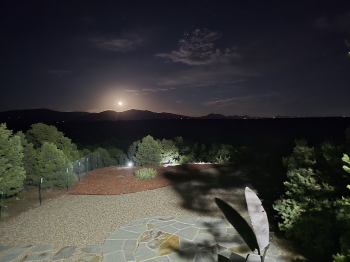 A moonlit backyard patio with flagstone, gravel, and evergreens overlooking distant mountains and scattered valley lights under a starry night sky.