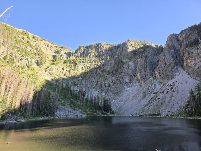 A calm alpine lake reflects steep rocky cliffs and forested slopes beneath a clear blue sky in a high mountain wilderness.