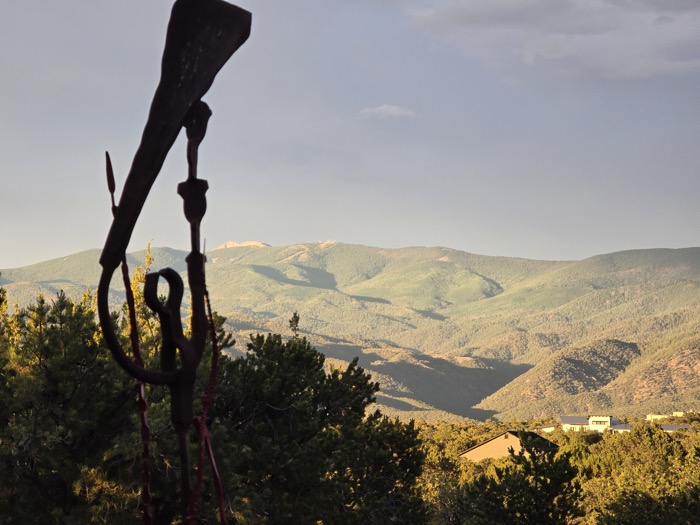 A silhouetted iron sculpture with hooked and spiral forms stands over sunlit green mountains, distant peaks, and valley rooftops at golden hour.