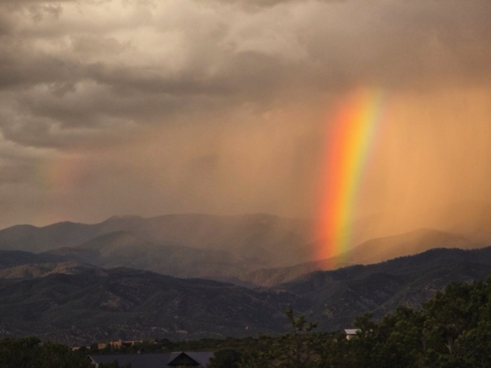 A bright double rainbow arches over layered mountains beneath storm clouds lit by warm orange sunset light.