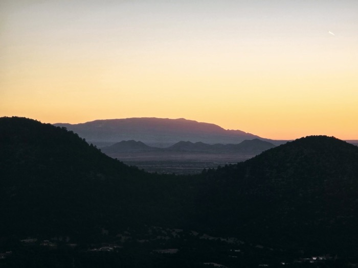 A broad flat-topped mesa glows pink against a warm sunset sky above a hazy valley with scattered lights and dark forested hills.
