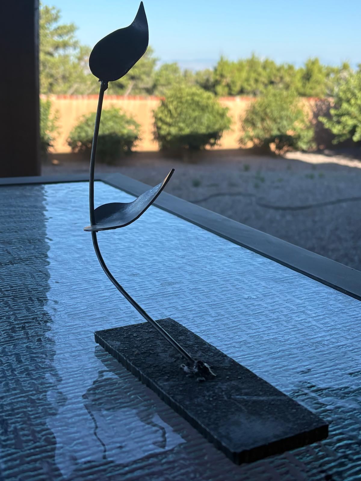 A slender black steel sculpture with two abstract leaf forms balanced along a curved stem on a rectangular base.