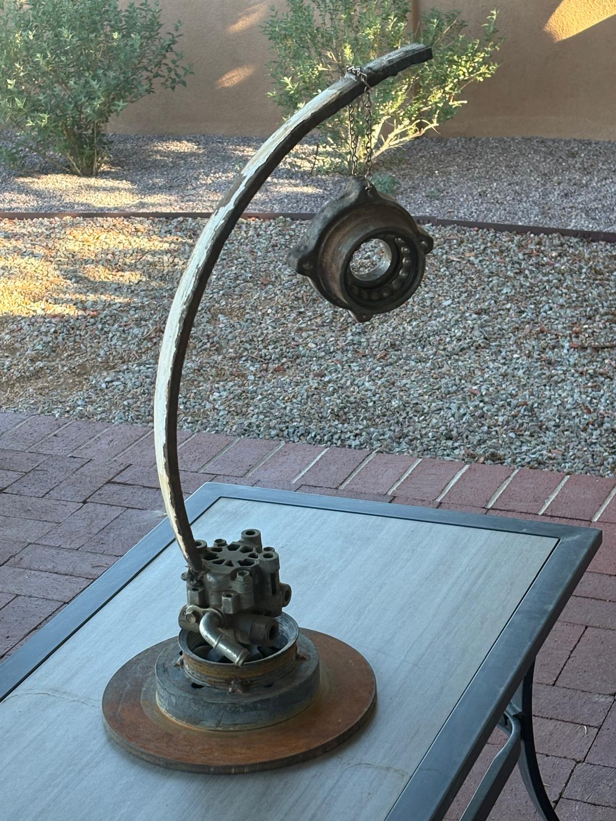 A welded steel sculpture with an arcing stem suspending a circular mechanical form above a base assembled from industrial components.
