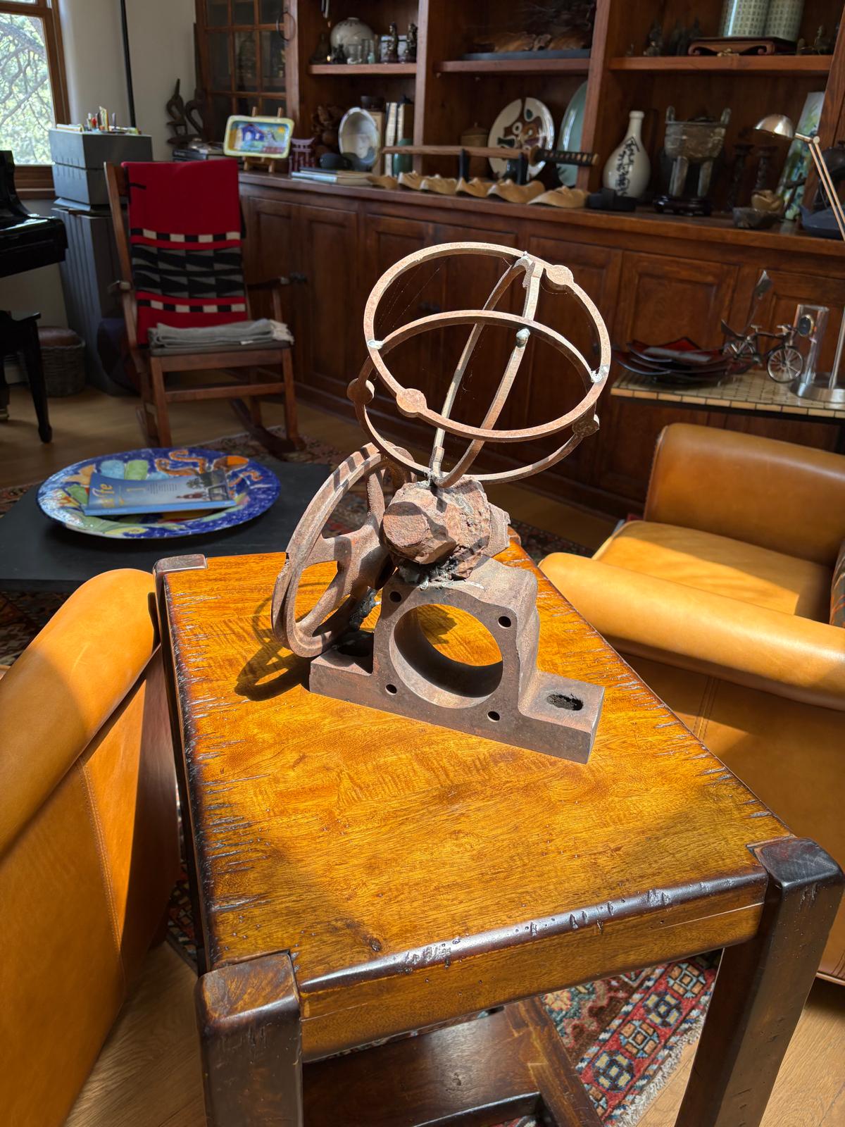 Front view of a rusted steel sculpture with intersecting orbital rings, curved cut-metal forms, stone, and an industrial base.
