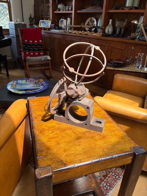 Front view of a rusted steel sculpture with intersecting orbital rings, curved cut-metal forms, stone, and an industrial base.