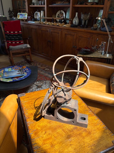 Side view of a rusted steel sculpture combining intersecting orbital rings, curved cut-metal forms, stone, and an industrial base.